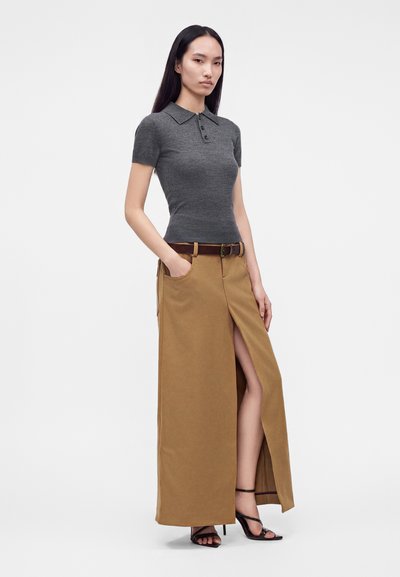 Femme portant un polo gris cintré à manches courtes, une longue jupe marron avec fente devant, une ceinture marron et des sandales noires à talons hauts, debout sur fond blanc.