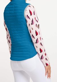 Blauer gesteppter Weste mit grauen Akzenten über einem langärmeligen, hellrosafarbenen Oberteil mit einem Muster aus roten, lila und braunen Blattformen.