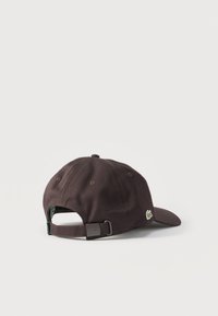 Casquette de baseball marron foncé avec sangle réglable et petit logo de crocodile vert sur le côté, posée sur un fond blanc.