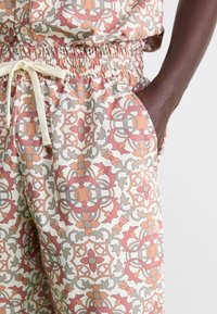 Pantalon à motifs avec un tissu léger et lisse présentant un design géométrique en rose, orange et gris, avec une taille élastique et des poches latérales.