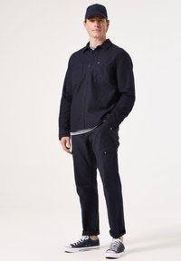 Marineblauwe overhemD met twee borstzakken, gecombineerd met marineblauwe cargobroeken en witte sneakers. Een bijpassende marineblauwe pet maakt de look compleet.