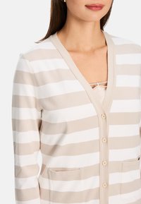 Beige- und weiß gestreifter Cardigan mit V-Ausschnitt, zwei vorderen Taschen und sechs Knöpfen auf der Vorderseite. Weiche Textur und lässige Passform.