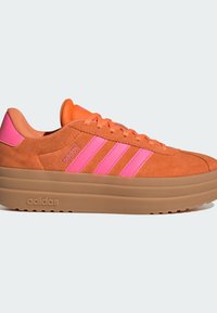 Oranje suède sneaker met een gelaagde beige platformzool, voorzien van felrode drie strepen en een logo op de zijkant. De veters zijn van dezelfde kleur als het bovenwerk.