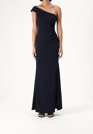 Kvinde iført en gulvlængde, marineblå, asymmetrisk aftenkjole med en højt skåret slids på låret og sorte åbne tå-hæle.