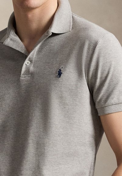 Vyras dėvintis šviesiai pilką trumpomis rankovėmis polo marškinėlį su tamsiai mėlynu siuvinėtu arklio ir raitelio logotipu ant kairiojo krūtinės.