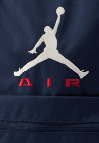 Το υφασμάτινο ύφασμα σε ναυτικό μπλε χρώμα διαθέτει μια μεγάλη λευκή κεντημένη σιλουέτα παίκτη μπάσκετ, με κόκκινο κείμενο "AIR" από κάτω. Το σχεδιασμένο κέντημα έχει υφή με ανάγλυφη επιφάνεια.