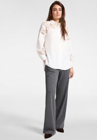 Vrouw staand, gekleed in een witte blouse met lange mouwen en kanten details, gecombineerd met grijze wijde broekspijpen en bruine puntschoenen.