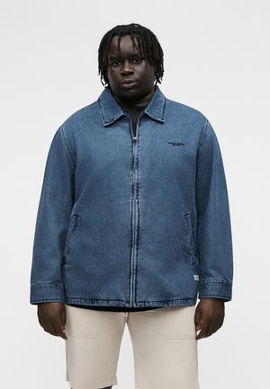 Férfi rasztahajjal, kék farmer zipzáras dzsekit visel fekete ing fölött, világos bézs rövidnadrágban, fehér háttér előtt állva.