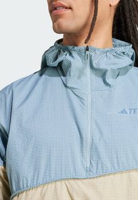 Suéter de tela escocesa azul con cuello con volantes, cierre de media cremallera y parte inferior beige. Presenta un sutil logo en el pecho.