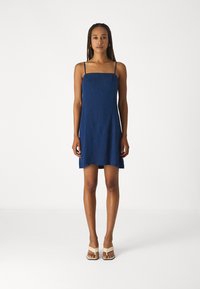 Robe sans manches bleu marine avec un décolleté carré, texture légère et ourlet évasé. Portée avec des sandales à talons beiges.