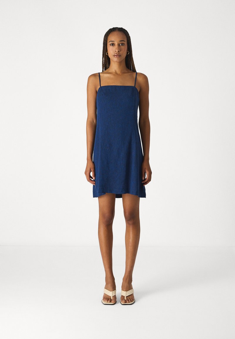 Robe sans manches bleu marine avec un décolleté carré, texture légère et ourlet évasé. Portée avec des sandales à talons beiges.