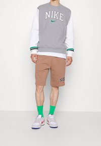 Man som bär en grå och vit Nike-tröja, bruna Nike-shorts, ljusgröna strumpor och vita sneakers med röda detaljer, stående mot en vit vägg.