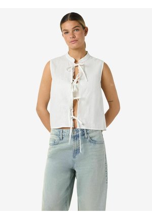 Young woman with pulled-back hair wearing sleeveless white tie-front blouse and light blue high-waist jeans, standing with hands behind back.