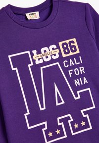 Sudadera púrpura con un estampado gráfico: "LOS ÁNGELES 86" y "CALIFORNIA" en blanco y crema. Cuello acanalado, tejido suave, detalle de cinco estrellas.