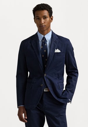 Blazer de marina con dos bolsillos delanteros, combinado con una camisa a rayas azul claro y una corbata con estampado. Pañuelo blanco en el bolsillo, cinturón marrón. Corte ajustado.