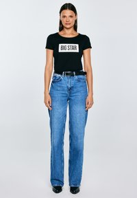 Kobieta stojąca przodem, ubrana w czarny t-shirt z napisem "BIG STAR", niebieskie jeansy z wysokim stanem, czarny pasek oraz czarne, spiczaste buty na szarym tle.