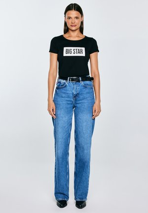 Moteris stovi veidu į priekį, dėvėdama juodą „BIG STAR“ marškinėlį, aukštakulnius mėlynus džinsus, juodą diržą ir juodus aštriapadžius batelius, pilkame fone.