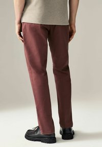 Pantalones burdeos con corte ajustado, fabricados en un tejido suave. Presentan bolsillos traseros y se combinan con zapatos negros de cordones y una camisa gris.