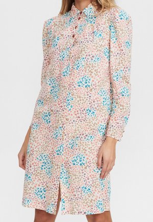 Femme portant une robe à fleurs mi-longue avec des petits motifs floraux bleus, roses et marron, devant boutonné et manches longues.