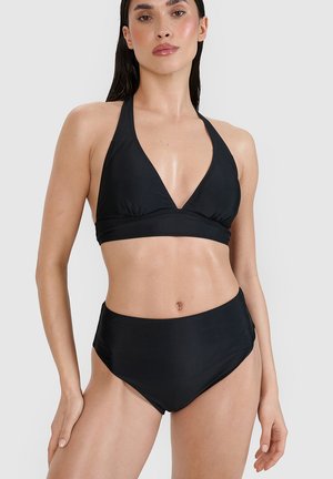 Kobieta w czarnym bikini na wiązanie za szyją i bikini z wysokim stanem stoi na tle jednolitego jasnego tła.