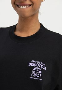 Close-up van een persoon die een zwart shirt draagt met een paarse discobal-afbeelding en tekst waarop staat: "Always Stay Groovy DISCOTECA Funky Way of Life."