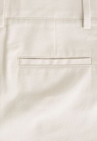 A világos bézs nadrág sima, texturált anyagból készült, hátul zsebes részlettel és övbújtatókkal, tiszta és szabott megjelenést kölcsönözve.