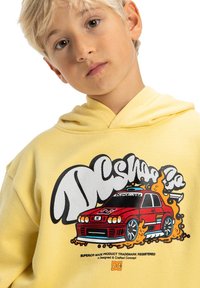 Blond dziecko w żółtej bluzie z kapturem z graficznym motywem czerwonego samochodu i stylizowanym białym napisem "DC Shoes" na przodzie.