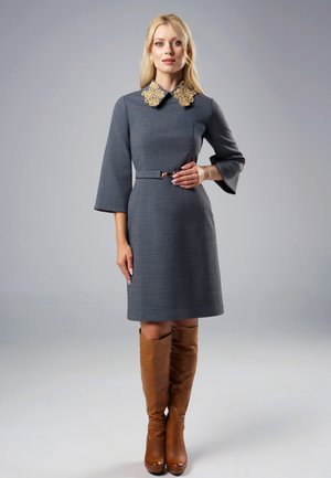 Kobieta ubrana w szarą sukienkę do kolan z ozdobnym złotym kołnierzem, rękawami w kształcie dzwonka, talią z paskiem oraz wysokimi brązowymi skórzanymi kozakami, stojąca nieruchomo.