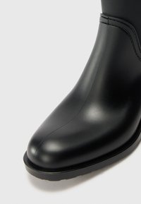 Bota de tornozelo preta em borracha com textura suave, bico pontiagudo e salto ligeiro, com costuras visíveis.