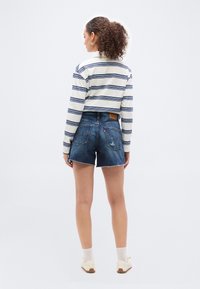 Γυναίκα με σγουρά μαλλιά που φοράει μακρυμάνικη μπλούζα λευκή με μπλε ρίγες, ξεφτισμένα τζιν σορτς, λευκές κάλτσες και παπούτσια χωρίς κορδόνια, γυρισμένη με την πλάτη.