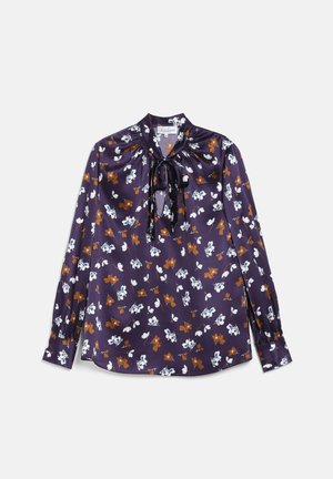Kvinde iført en lilla blomster-satinbluse med sløjfe, blå højttaljede vidde jeans, sort bælte, sorte loafers og guldsmykker.