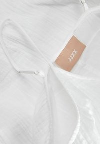 Tela blanca con una superficie texturizada y arrugada. Presenta una etiqueta beige con "JXX" impresa en blanco. Incluye tirantes delgados en la parte superior.