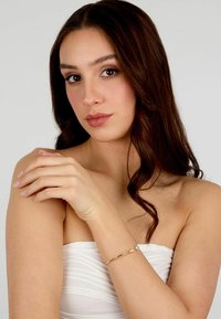 Braccialetto rigido dorato con un accento di pietra preziosa trasparente, indossato su un braccio nudo su uno sfondo chiaro. Design minimale e liscio.