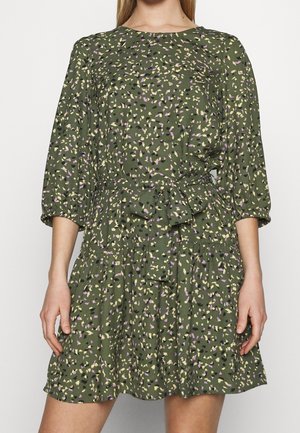 Frau trägt ein olivgrünes Kleid mit kleinem gelbem, schwarzem und lila Blumenmuster, dreiviertel Ärmeln und einer Schleife zum Binden an der Taille.