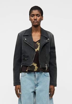 Γυναίκα που φοράει μαύρο τζιν μπουφάν cropped πάνω από μπλούζα με σκούρο λουλουδάτο σχέδιο και ανοιχτόχρωμο τζιν με ψηλή μέση, στέκεται μπροστά σε απλό φόντο.