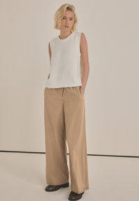 Weißes ärmelloses Strickoberteil mit geripptem Halsauschnitt, kombiniert mit beigen, gestreiften, weiten Hosen. Das Model posiert mit den Händen in den Taschen.