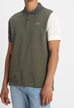 Homme portant un polo bicolore avec un devant vert olive, un col et une manche gauche gris foncé, et une manche droite blanche, assorti à un jean bleu clair.
