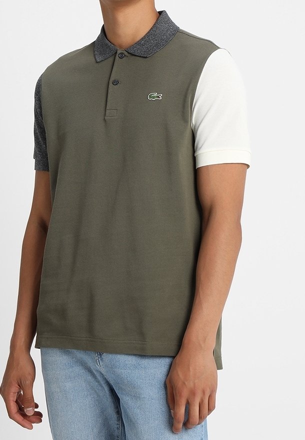 Homme portant un polo bicolore avec un devant vert olive, un col et une manche gauche gris foncé, et une manche droite blanche, assorti à un jean bleu clair.