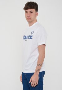 T-shirt bianco in cotone con un grande logo blu sul davanti. Design a girocollo con maniche corte. Forma semplice e classica.