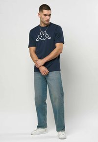 Mann iført marineblå t-skjorte med logo, lyseblå jeans og hvite joggesko står mot en ensfarget hvit bakgrunn.