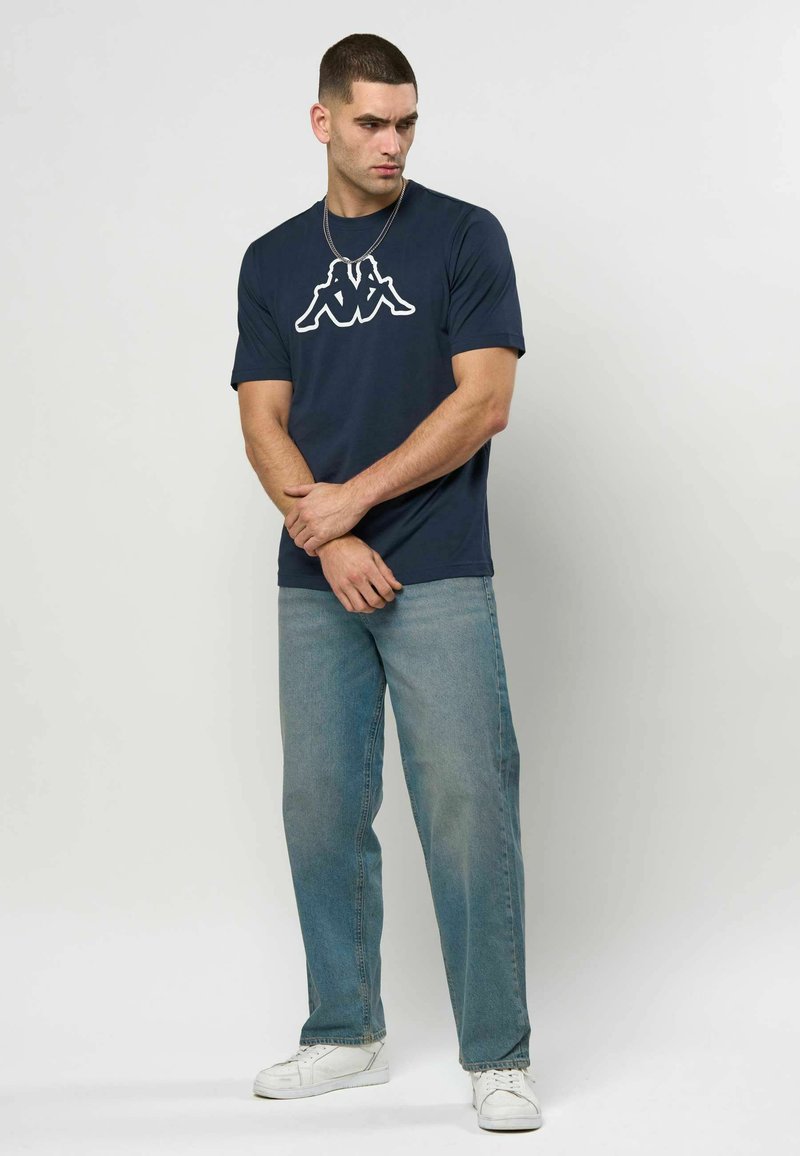 Mann iført marineblå t-skjorte med logo, lyseblå jeans og hvite joggesko står mot en ensfarget hvit bakgrunn.