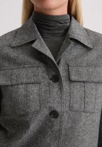 Camicia grigia con colletto, dotata di due tasche sul petto e di un tessuto intrecciato e strutturato. La camicia è indossata sopra un dolcevita scuro.