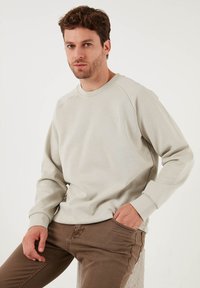 Felpa grigia chiara con collo tondo, polsini a costine e logo discreto, abbinata a pantaloni marroni. Tessuto morbido, vestibilità comoda, design semplice.