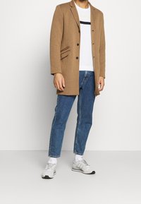 Brązowy wełniany płaszcz z bocznymi kieszeniami, sześć czarnych guzików, noszony na białym swetrze w poziome paski, zestawiony z niebieskimi dżinsami i szarymi tenisówkami.