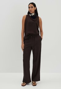 Bruine mouwloze top met een gedrapeerde halslijn, gecombineerd met losse bruine broek met een elastische tailleband en zijzakken. Zwarte slippers.