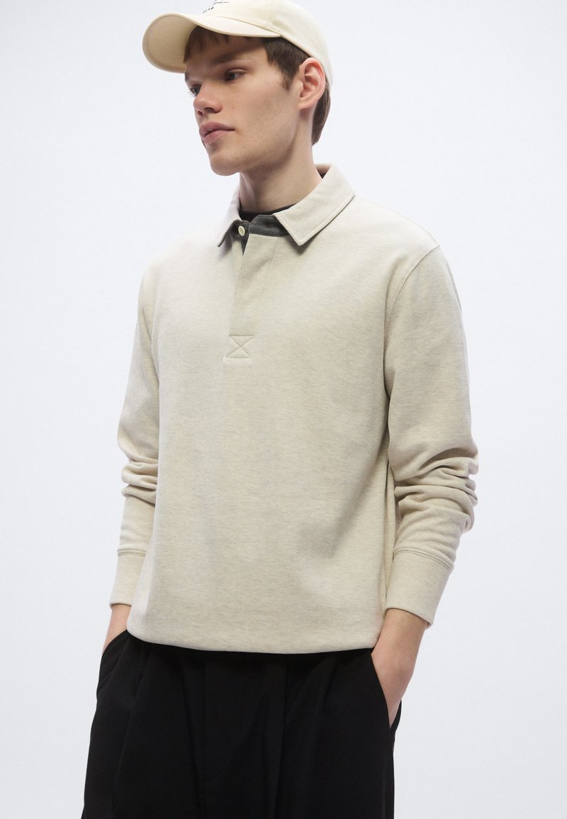 Beige long-sleeve polo shirt with a collar, featuring a textured front placket and a relaxed fit. Worn with black trousers and a light cap.