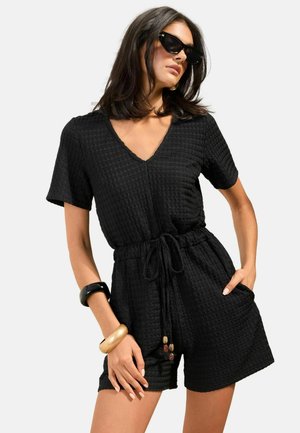 Kvinde iført sort, tekstureret kortærmet heldragt med snøre i taljen, accessoriseret med sorte solbriller og guld- og sorte armbånd.