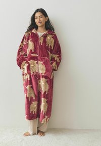 Red plush hooded robe featuring tan sheep and cow patterns, long sleeves, side pockets, and a front zipper. Worn over beige pants.