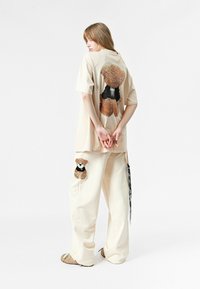 Camisa beige con un gráfico texturizado de un oso de peluche en la parte posterior; pantalones beige a juego con un parche más pequeño de un oso de peluche en el lateral.