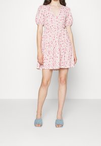 Mujer vistiendo un vestido corto de flores en tonos rosa y blanco con mangas abullonadas y tacones destalonados de color azul claro, de pie contra un fondo liso.